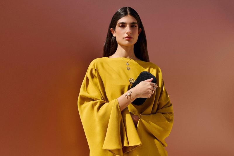 Elegant woman in mustard dress holds a black purse against a gradient backdrop. Fashion, jewelry, and studio lighting create a sophisticated mood.