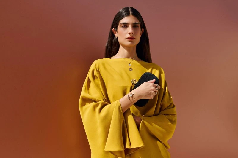 Elegant woman in mustard dress holds a black purse against a gradient backdrop. Fashion, jewelry, and studio lighting create a sophisticated mood.