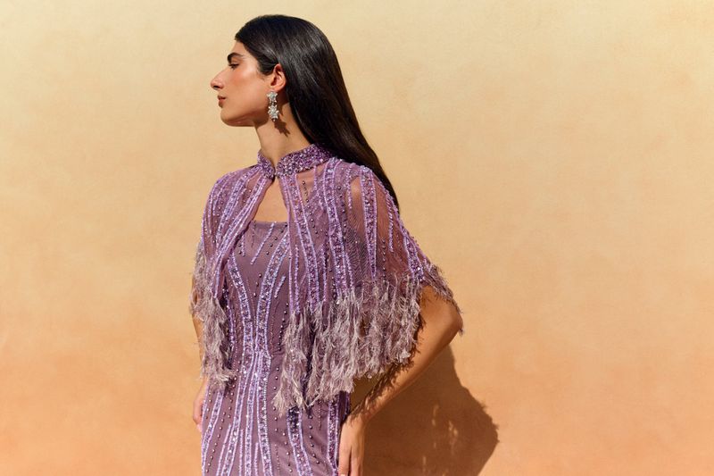 Brunette woman poses in beaded and feathered, pale purple gown and cape in front of a neutral colored wall in a medium shot.