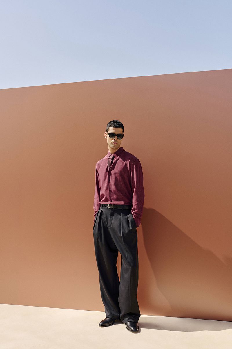 A stylish adult male stands in a fashionable burgundy button-down shirt and black slacks in front of a peach-colored wall outdoors.
