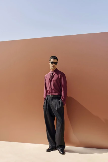 A stylish adult male stands in a fashionable burgundy button-down shirt and black slacks in front of a peach-colored wall outdoors.