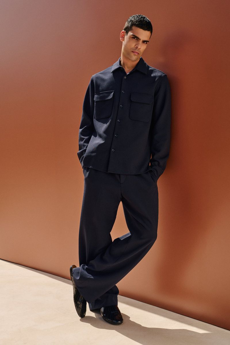 Portrait of a young adult man leaning against an orange wall, wearing a dark blue shirt and pants with dark shoes. Studio shot.