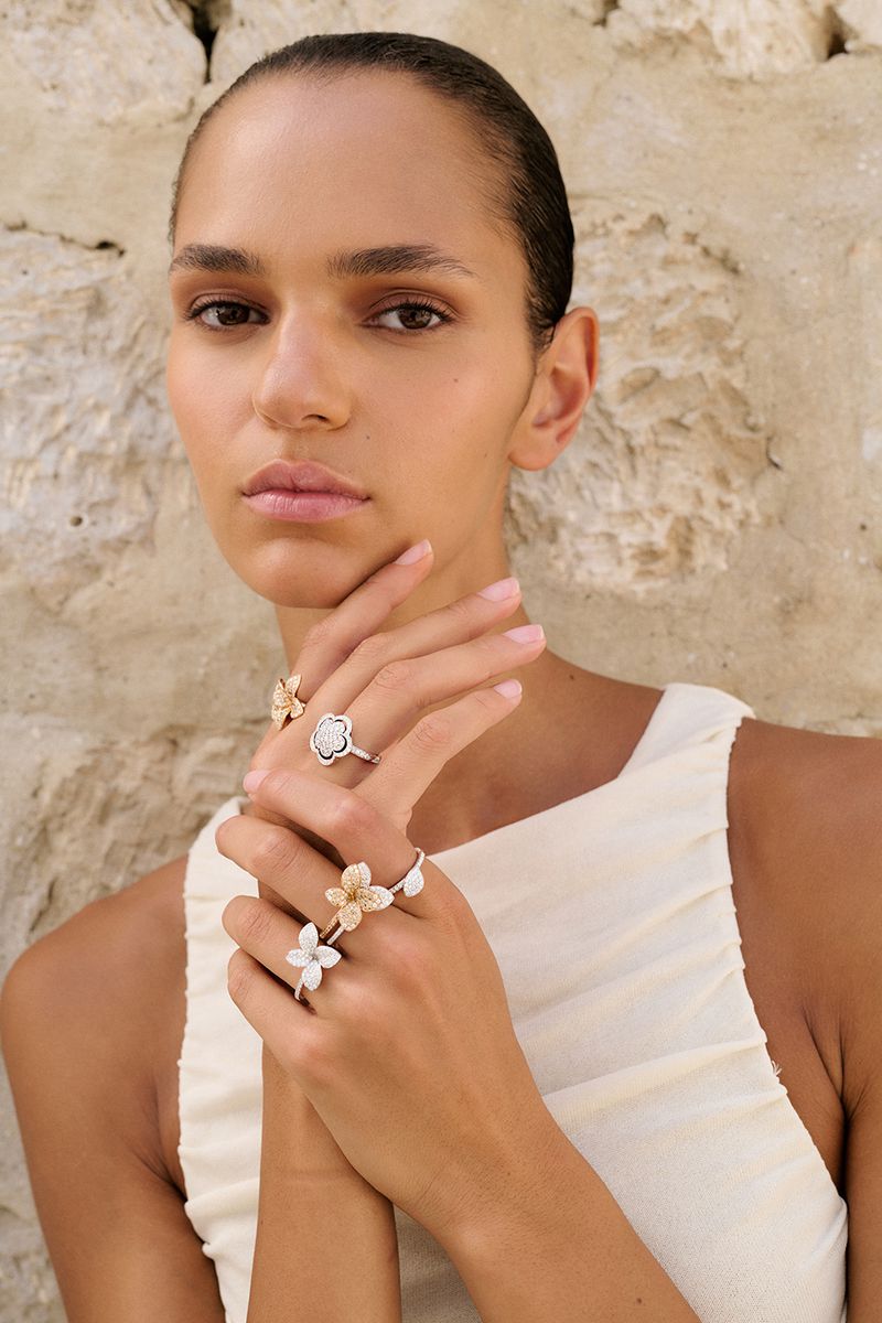 Elegant woman poses wearing flower-shaped rings against a textured wall backdrop in a close-up beauty shot showcasing jewelry.