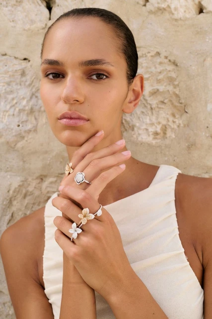 Elegant woman poses wearing flower-shaped rings against a textured wall backdrop in a close-up beauty shot showcasing jewelry.
