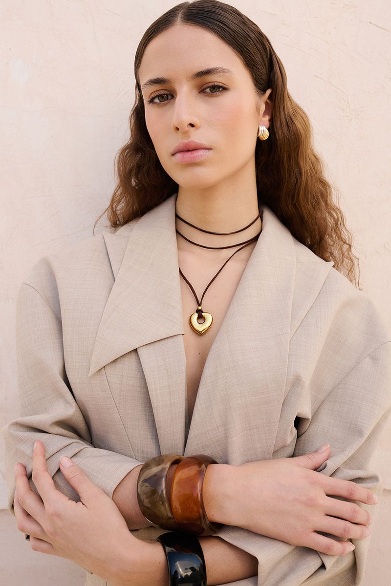 Woman in a tan jacket poses with gold heart necklace and stacked bangles against a neutral background in a medium portrait shot.