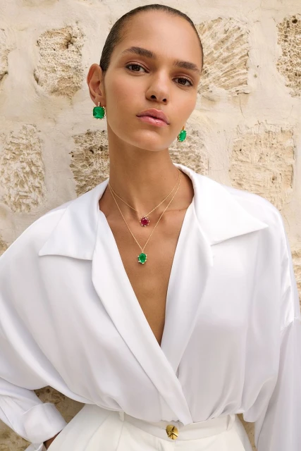 Portrait of a woman modeling jewelry, wearing emerald and ruby pendants and earrings with a stone wall visible in the background.