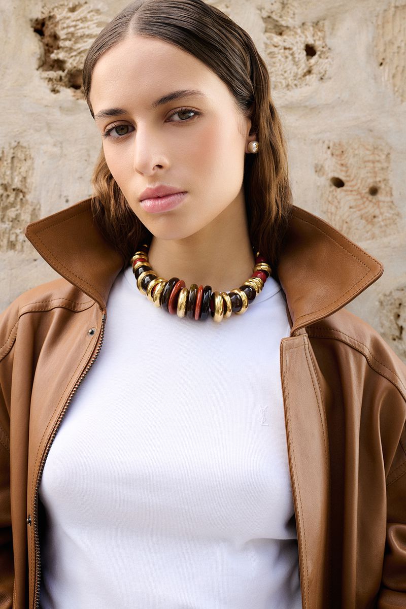 Close-up portrait of a woman wearing a white top, brown leather jacket, and chunky statement necklace against a stone wall.