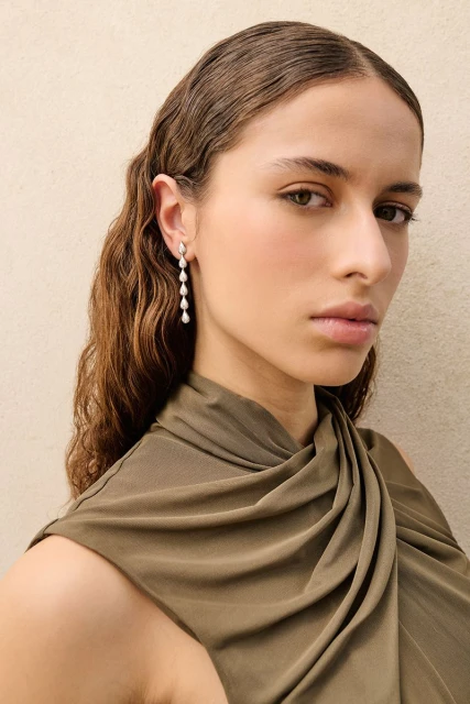 Portrait of a woman with wavy brown hair wearing teardrop earrings and a draped green dress against a neutral background.