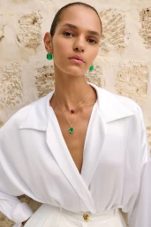 Model Chiara Bimbatti wearing emerald earrings and gemstone necklaces with a white shirt against a textured stone wall