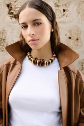 Close-up portrait of a woman wearing a white top, brown leather jacket, and chunky statement necklace against a stone wall.