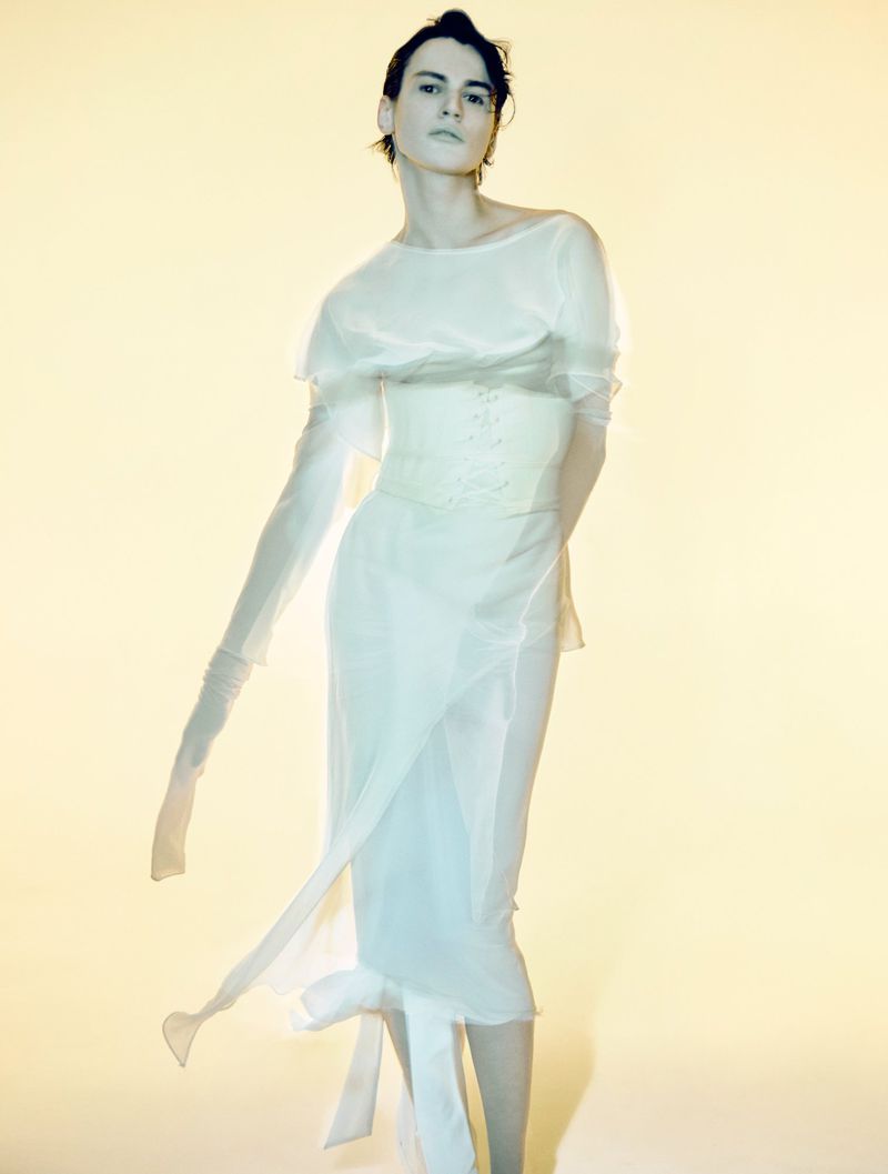 A slender, fair-skinned person poses in a long, flowing white dress in a studio setting with soft lighting and a light-yellow background.