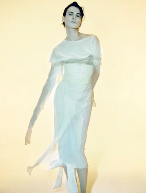 A slender, fair-skinned person poses in a long, flowing white dress in a studio setting with soft lighting and a light-yellow background.