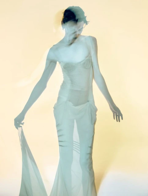 Ghostly woman in dress poses in photo studio with slow shutter speed, creating motion blur and ethereal effect against solid background.