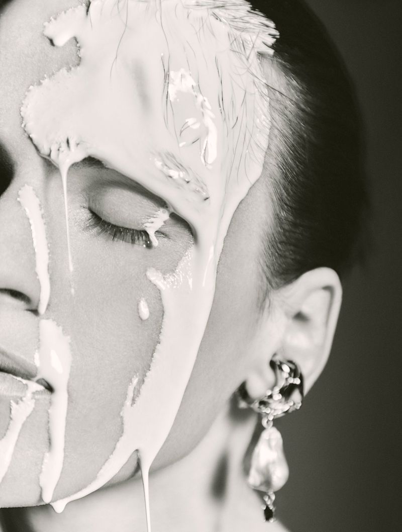 Close-up portrait of a woman with white cream makeup dripping down her face, one eye closed, wearing dangling pearl earring.
