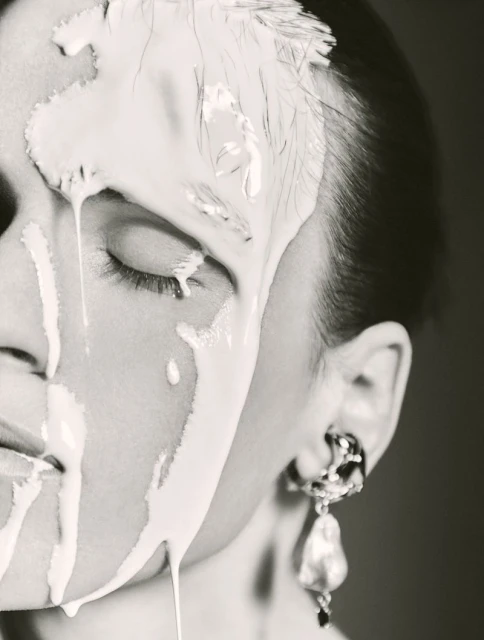 Close-up portrait of a woman with white cream makeup dripping down her face, one eye closed, wearing dangling pearl earring.