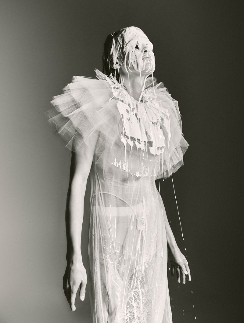 Adult female poses in a semi-sheer dress, covered in white liquid against a grey background, showcasing fashion and visual art.