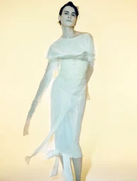 Model in a sheer white dress with flowing fabric against a soft yellowish background