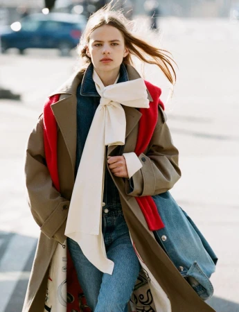 Model in camel trench coat and denim wearing a white bow scarf outdoors in the city for Harper's Bazaar France
