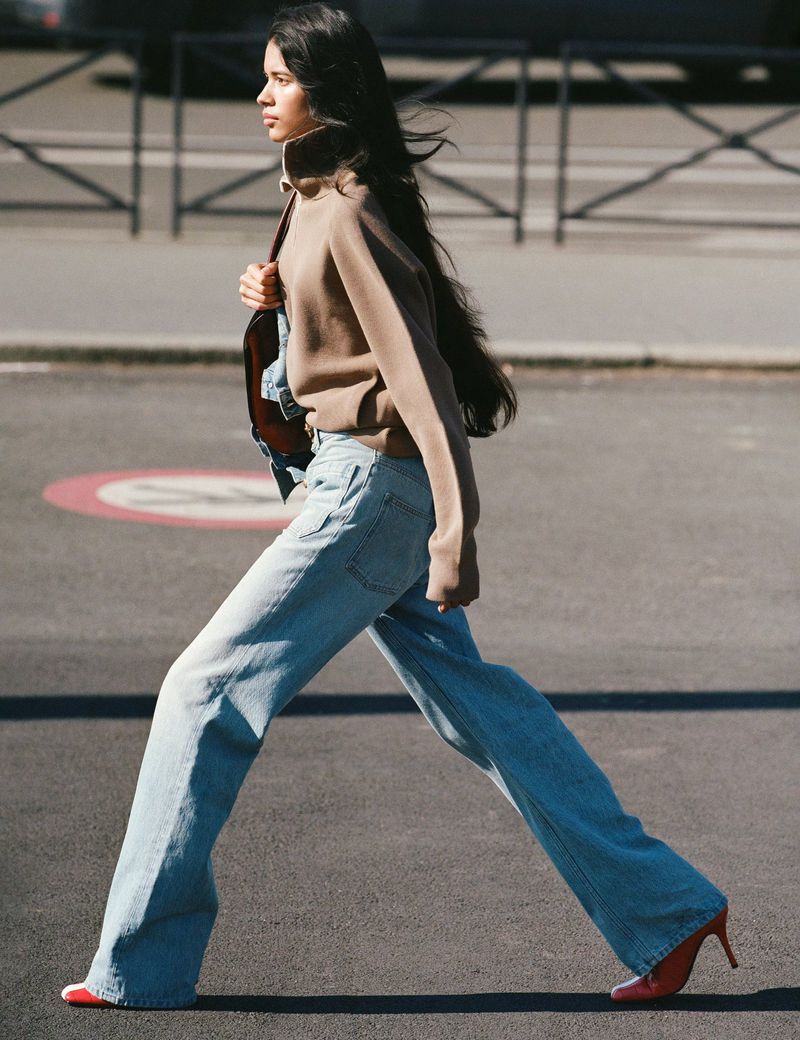 Woman in a brown turtleneck sweater and wide-leg jeans walks on a city street with red high heels and a shoulder bag. 