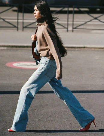 Woman in a brown turtleneck sweater and wide-leg jeans walks on a city street with red high heels and a shoulder bag. 