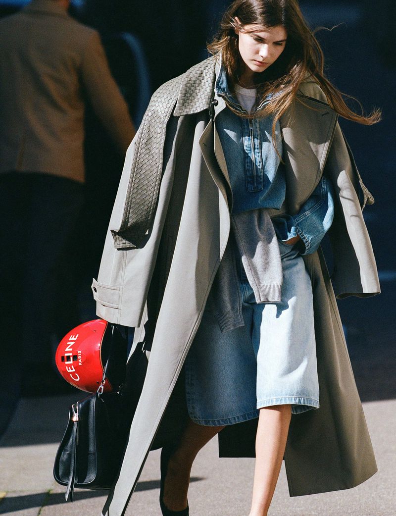 Young woman in a denim outfit and grey trench coat walking outside holding a red helmet and black handbag in a fashion editorial.