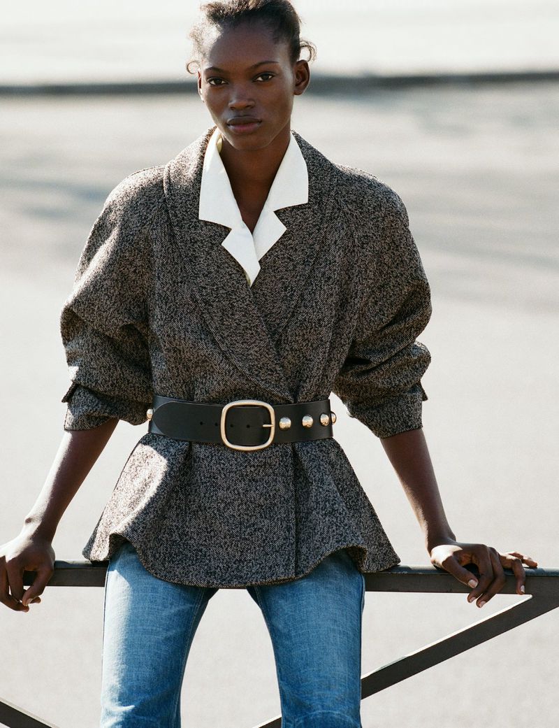 Model wearing a textured grey jacket, white collar, and wide black belt leaning against an outdoor metal railing.