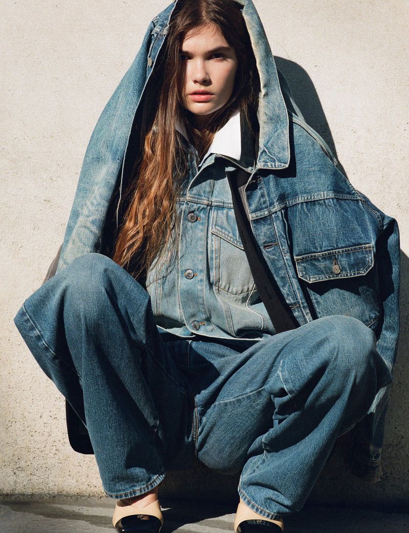 Adult female in denim jacket and jeans crouching against a concrete wall. Denim jacket is oversized with a hood. Neutral lighting.
