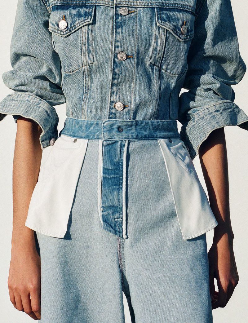 A cropped studio shot of a person wearing a denim jacket and light wash denim pants with white fabric accents, showcasing a fashion-forward design.