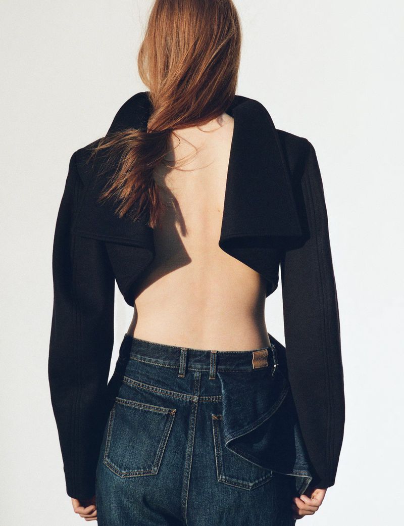 Back view of a woman with auburn hair wearing a black crop top with an open back and dark blue denim jeans in a white studio.