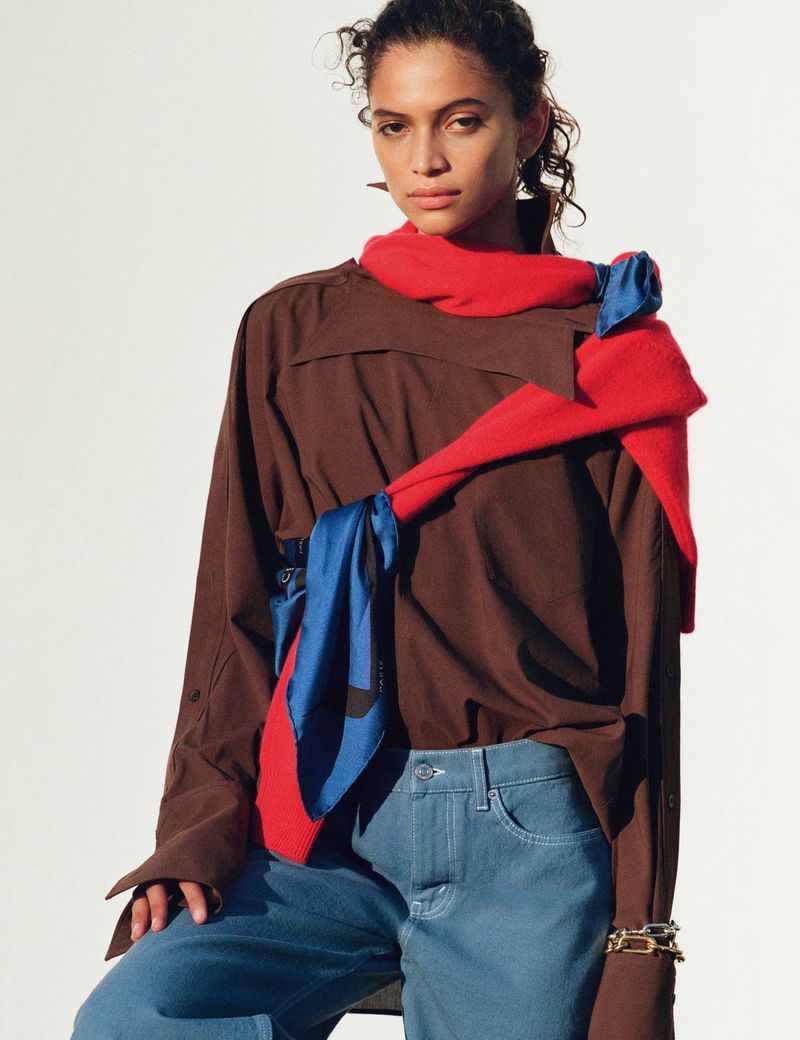 Fashion shot of a woman in blue jeans and brown button-down shirt, with a blue and red scarf, posing in a brightly lit indoor setting.