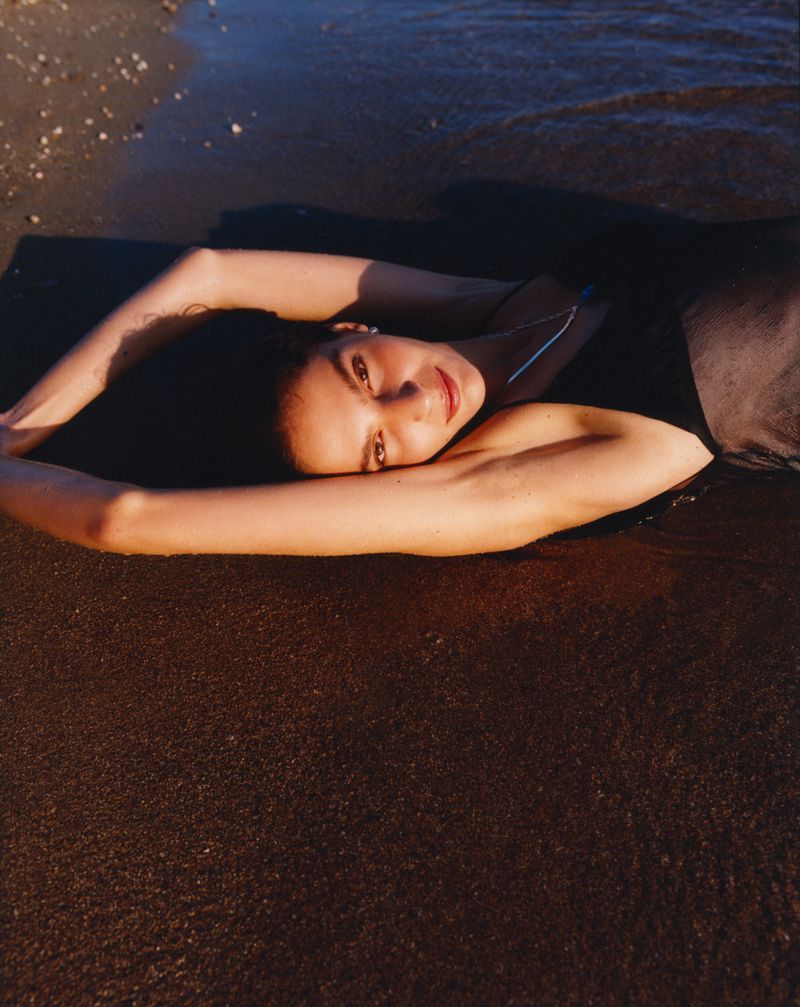 A woman lies on a sandy beach at the edge of the water. She is smiling with arms outstretched in the warm sunlight.