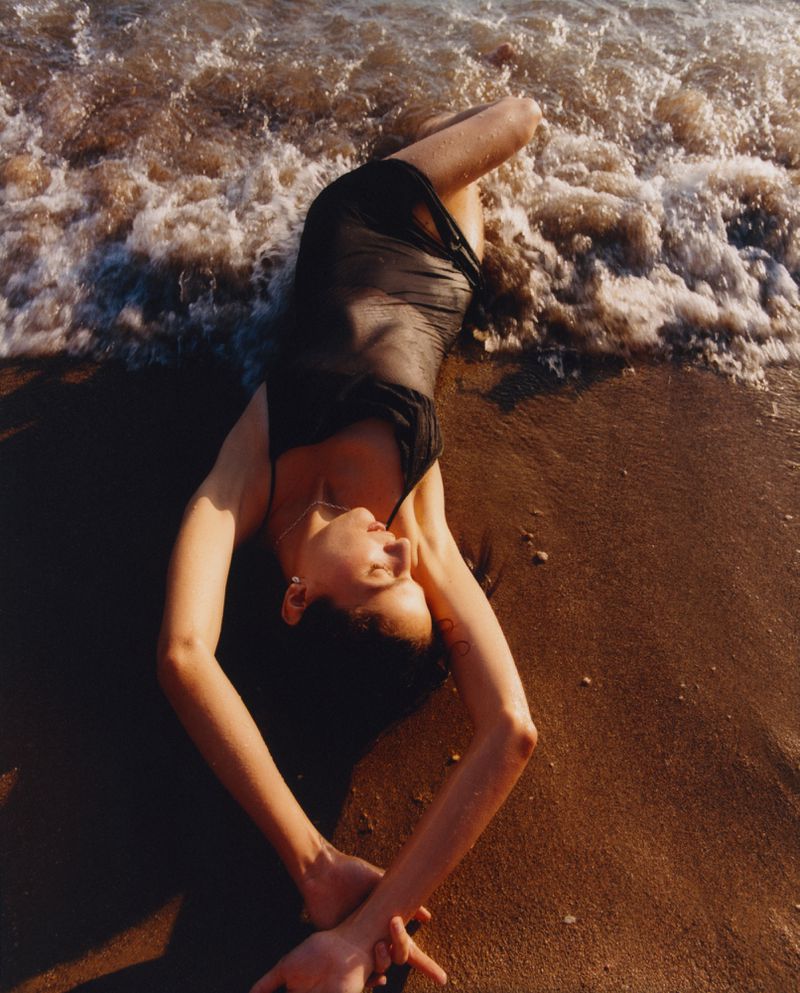 Woman in a black dress lying on a sandy beach with waves lapping nearby, viewed from an overhead angle on a sunny day.