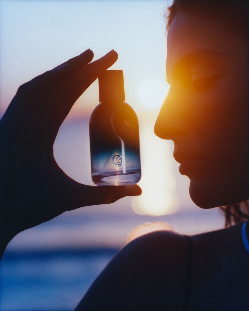 Silhouette of a woman holding a perfume bottle at sunset, with orange and blue sky and sea in the background, and her hand partially obscuring the bottle.