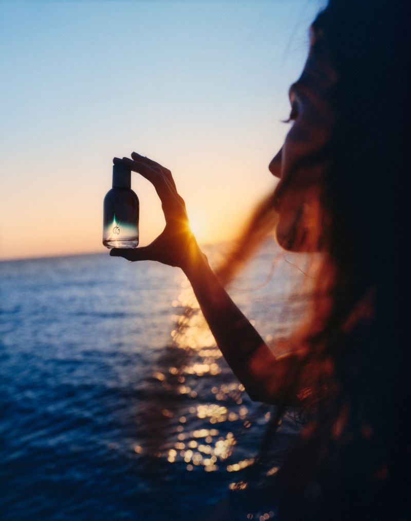 Woman holds a clear bottle of liquid against a backdrop of ocean water and sunset, creating a serene and visually striking image.
