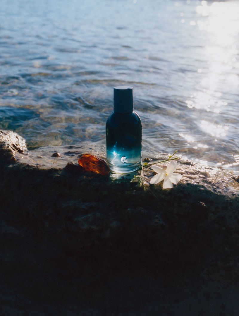 Product shot of a blue fragrance bottle with a black cap resting on a stone surface beside a translucent brown stone and white flower. 