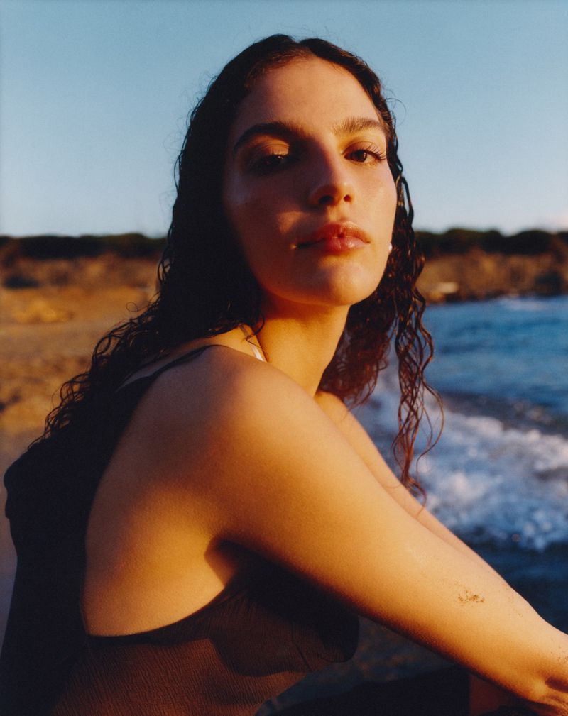 Adult woman poses at the beach in a close-up shot. The model has long curly hair, tan skin, and wears a dark top on a sunny day.