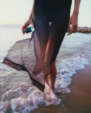 A woman in a black dress stands in the surf on a sandy beach, holding a blue bottle with water. The scene is bathed in soft light.