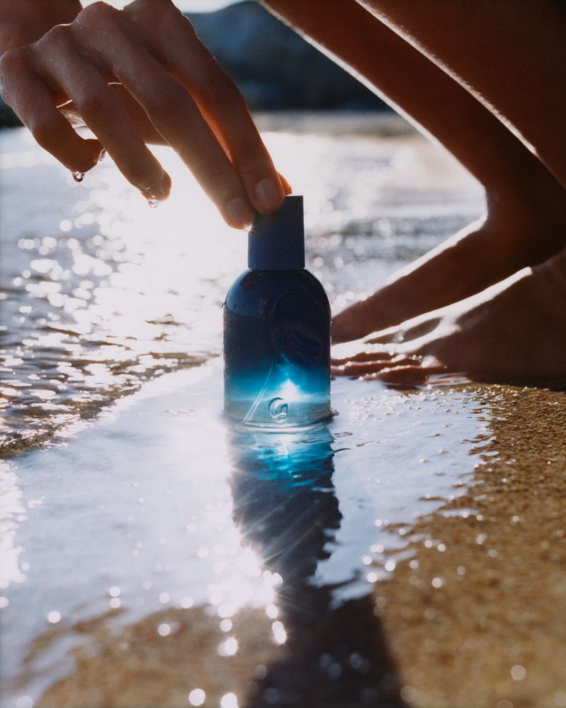 Close-up of a blue-toned perfume bottle in sand, hands near cap, shallow water, sunny outdoor beach setting.