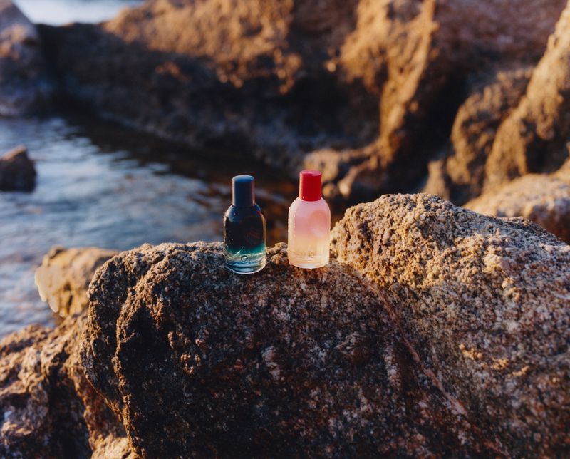Two perfume bottles sit on a large rock by the ocean. One has a blue cap and contains green liquid, the other, a red cap with pink liquid.