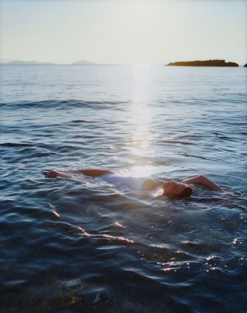 Woman in white swimsuit floats peacefully in the rippling sea under the bright sun with a small, rocky island in the distance.