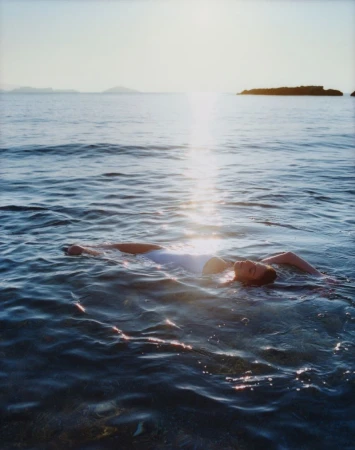 Woman in white swimsuit floats peacefully in the rippling sea under the bright sun with a small, rocky island in the distance.