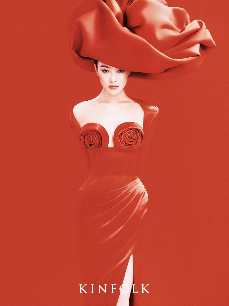 Elegant woman in a red dress and large red hat poses against a red background in a fashion editorial shot with soft, diffused lighting.