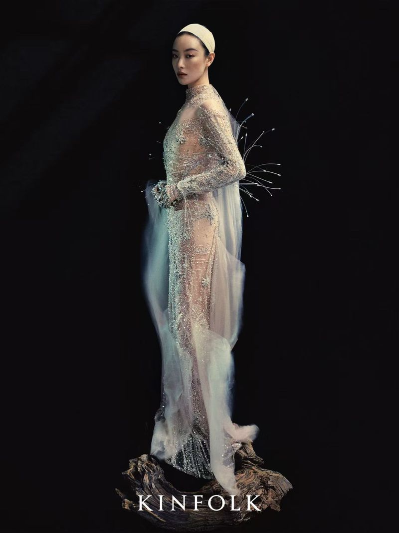 Elegant woman in beaded gown with a sheer cape, standing on a wooden base against a dark background, fashionably posed, looking at the camera.