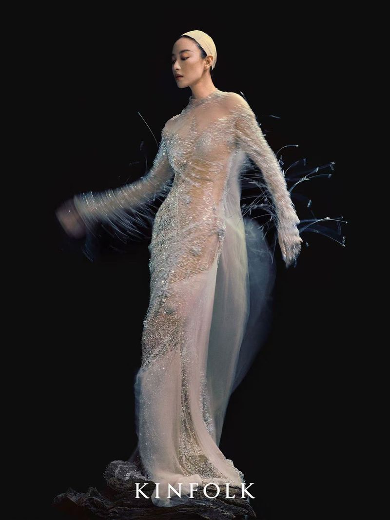 A woman in an embellished gown strikes a pose in front of a stark black background. The word 'KINFOLK' is visible at the bottom.