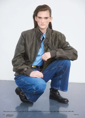 Person squats wearing a leather jacket, light blue shirt, jeans, and black boots, in a fashion-style portrait on a white background.