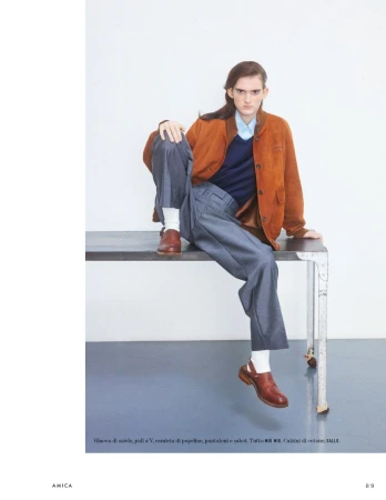 A person with long brown hair, wearing a rust-colored suede jacket, navy sweater, blue shirt, and grey trousers, sits on a metal table.