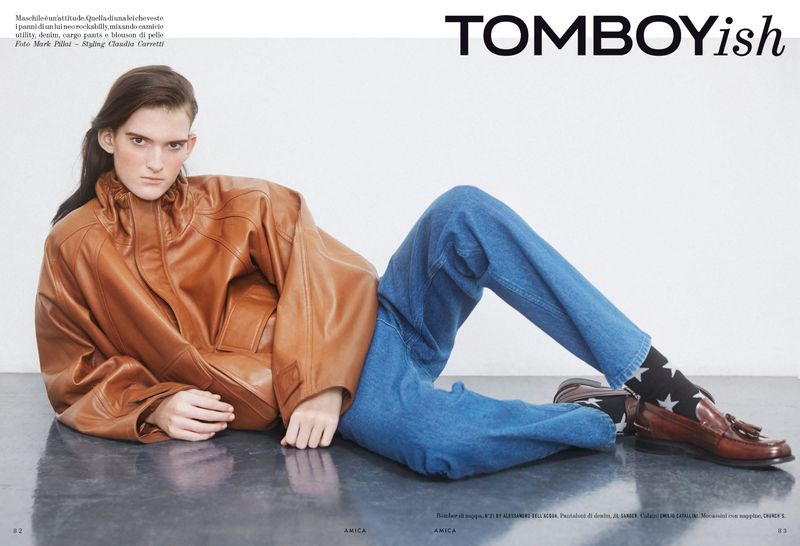 A model lying on a gray floor against a white wall, wearing a brown leather jacket, blue jeans, and star-patterned socks with loafers.