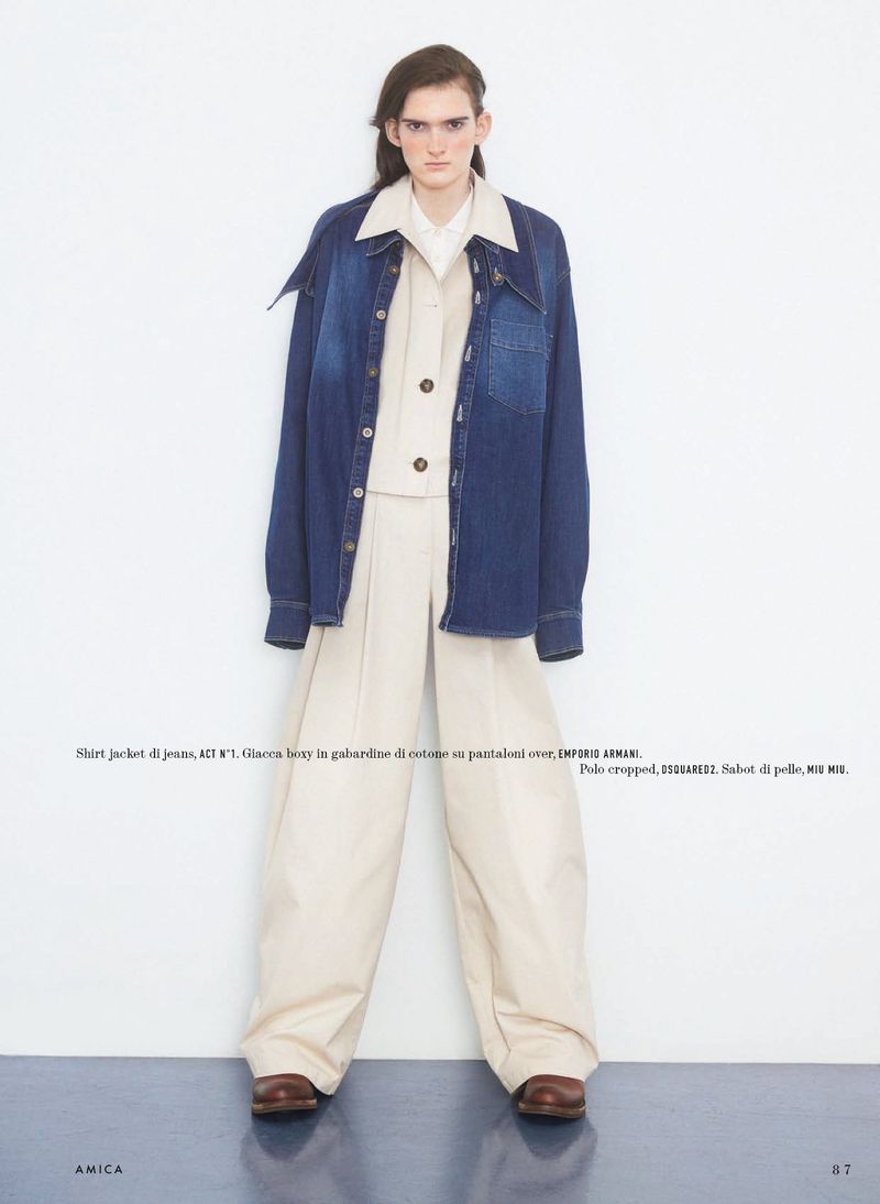Fashion model poses in oversized denim jacket, button-down shirt, and wide-legged beige pants in a studio setting, fashion editorial.
