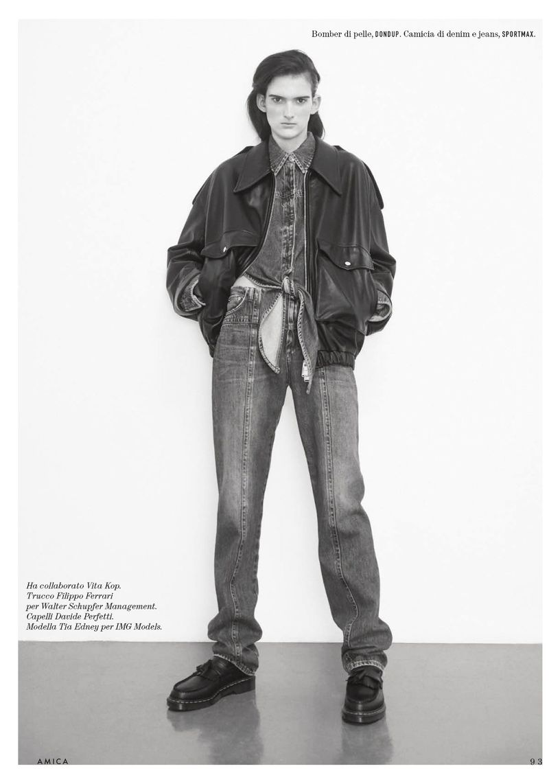 Full-length fashion shot: slender figure posing in denim shirt, jeans, and oversized jacket against a stark white backdrop.