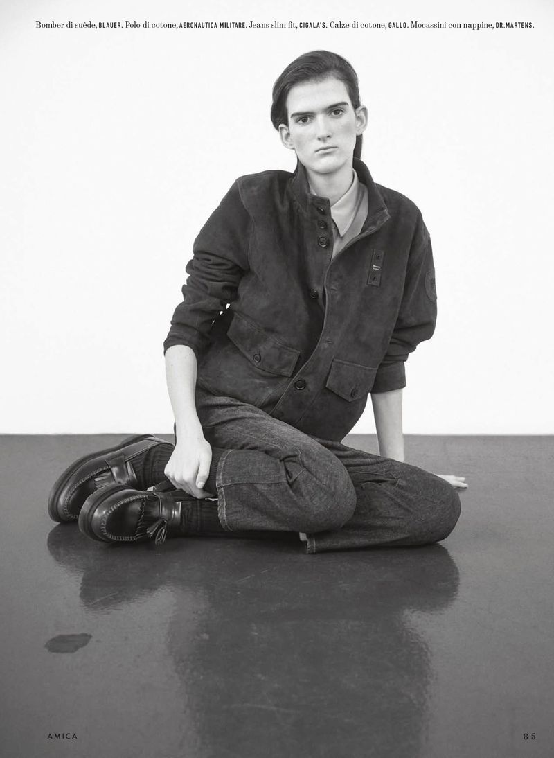 Fashion shot of a person seated in jeans and a jacket, with a neutral expression against a plain backdrop and sleek studio flooring.